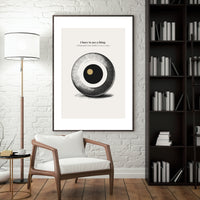 Framed artwork on a wall above a chair in a room with a lamp and bookshelf.