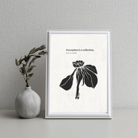 Framed artwork with black floral design and text on a white background, placed next to a vase with greenery on a textured wall.