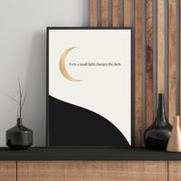 Framed artwork with a gold crescent moon and text on a white background, displayed on a wooden shelf.